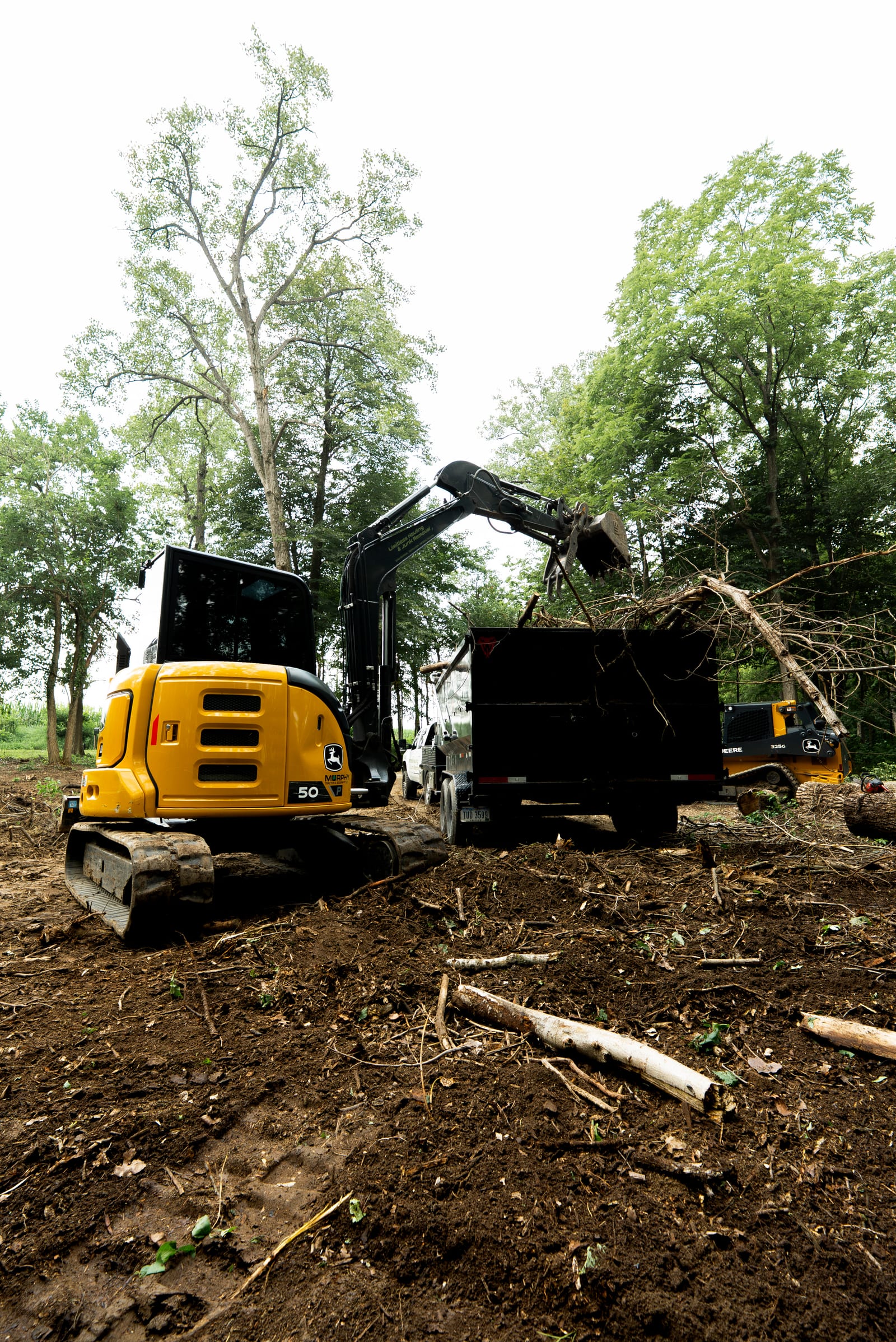 An excavator with a grapple loading cut brush and timber into a dump trailer